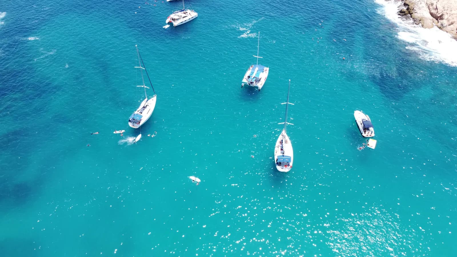 An aerial view of sailboats and motorboats anchored in bright turquoise water beside a rocky shore, with swimmers nearby.