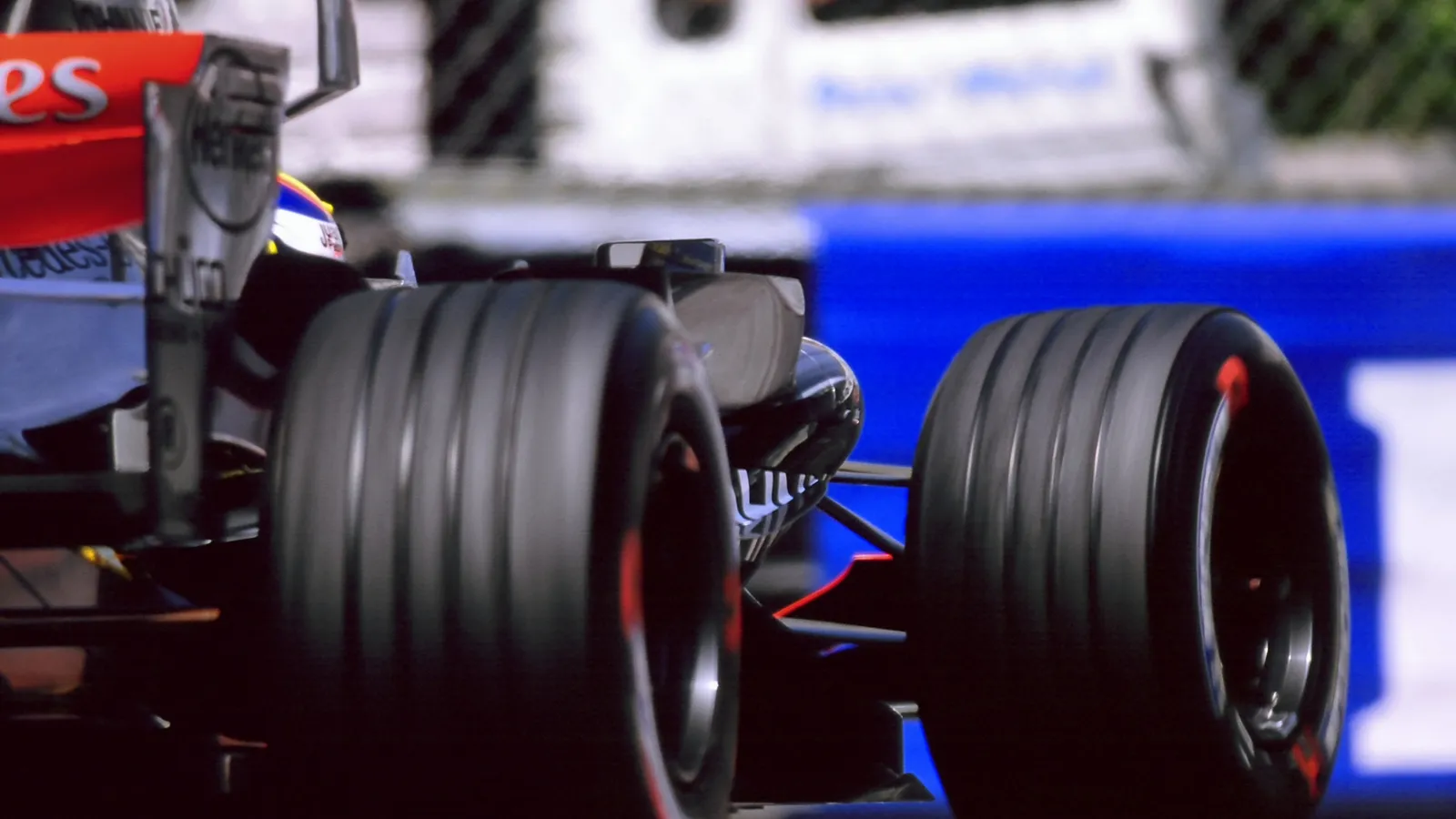 The rear of a Formula 1 car at speed, its wide rear tyres and rear wing in sharp focus against a blurred blue trackside barrier.