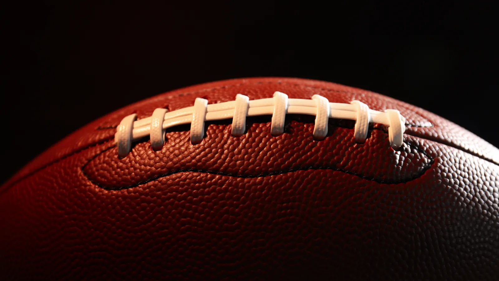 A close-up of an American football's white laces and pebbled leather against a dark background.